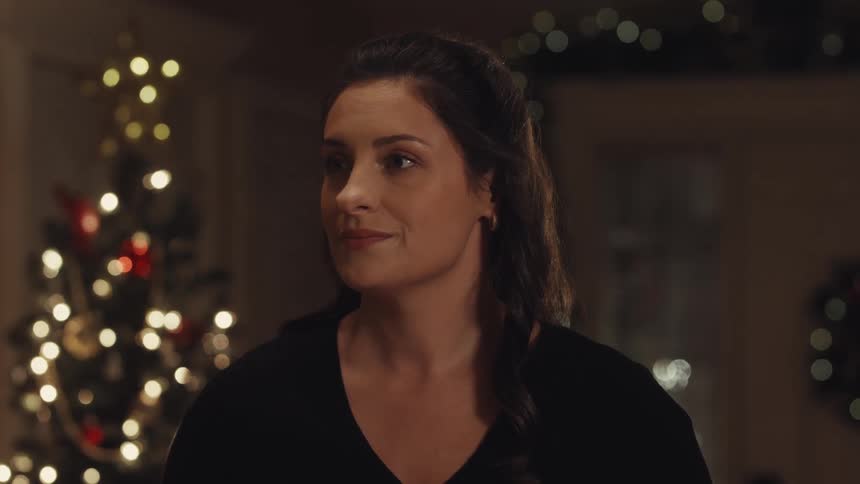 A woman sits indoors looking toward someone beside a glowing christmas tree