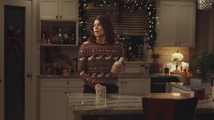 Woman in christmas sweater pours eggnog into mug in decorated kitchen