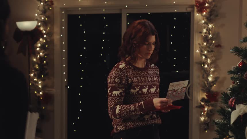 A woman wearing a brown patterned sweater stands in front of a decorated tree and sparkling lights as she opens a red envelope to read a card and smiles before handing it to a person off screen