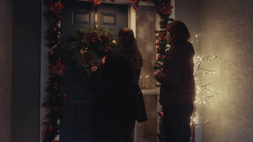 Two women talk at the doorway of a house decorated with christmas lights and garlands