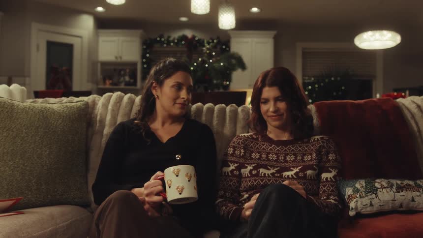 Two women sit on a sofa in a festive cozy living room sharing a mug and a warm conversationas the video progresses they lean into each other and break into shared laughter while drinking their beverage