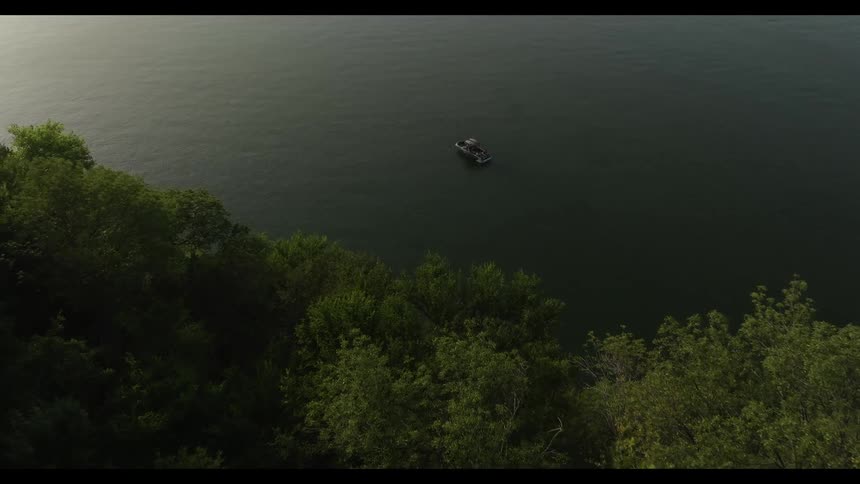 A wide view from above shows a small boat floating on the calm dark water of a large lake next to a steep shoreline covered in dense green trees during the dim light of dusk