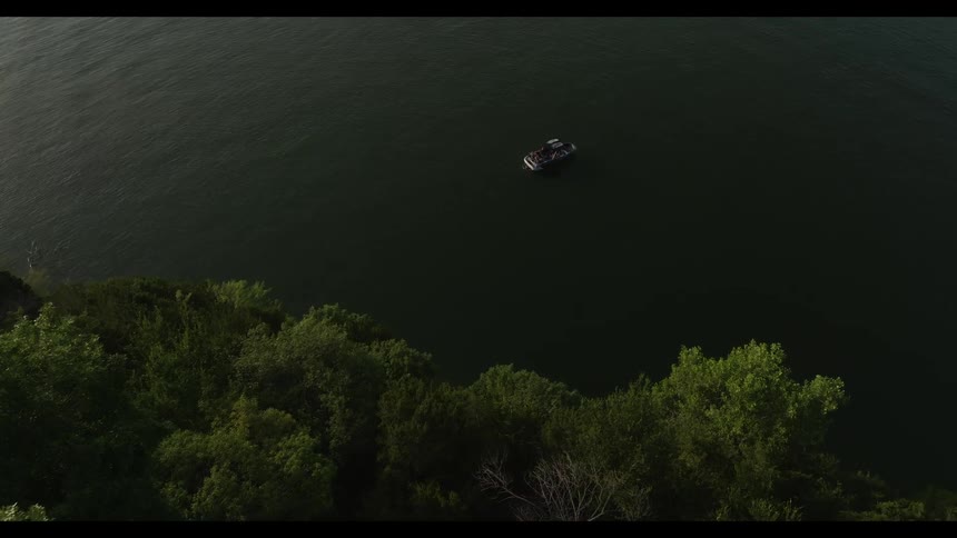 An aerial drone shot captures a small boat floating calmly on a dark body of water near a lush green shorelinethe camera slowly pans over the dense treetops of a cliffside revealing the vast expanse of the lake below
