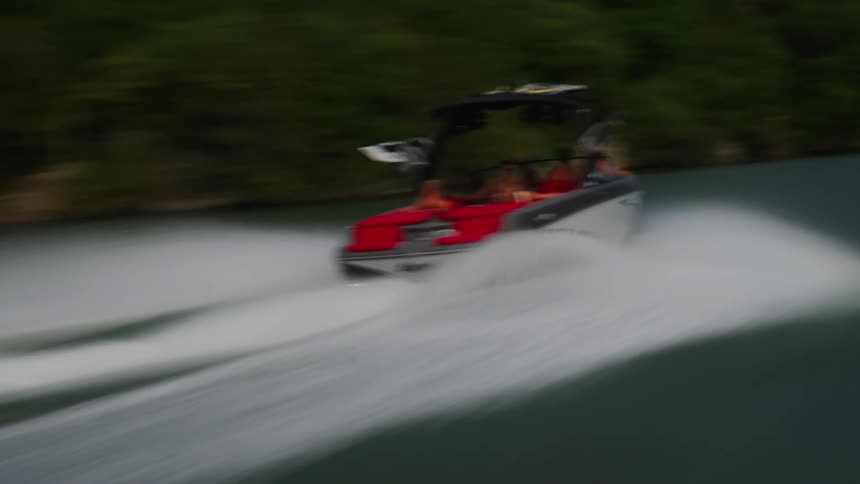 A red and grey motorboat carrying a group of friends speeds across a calm lake near a green shoreline and rocky cliffs during a bright day