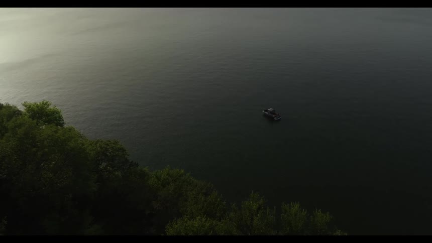 A wide view from above shows a small boat floating on the calm dark water of a large lake next to a steep shoreline covered in dense green trees during the dim light of dusk
