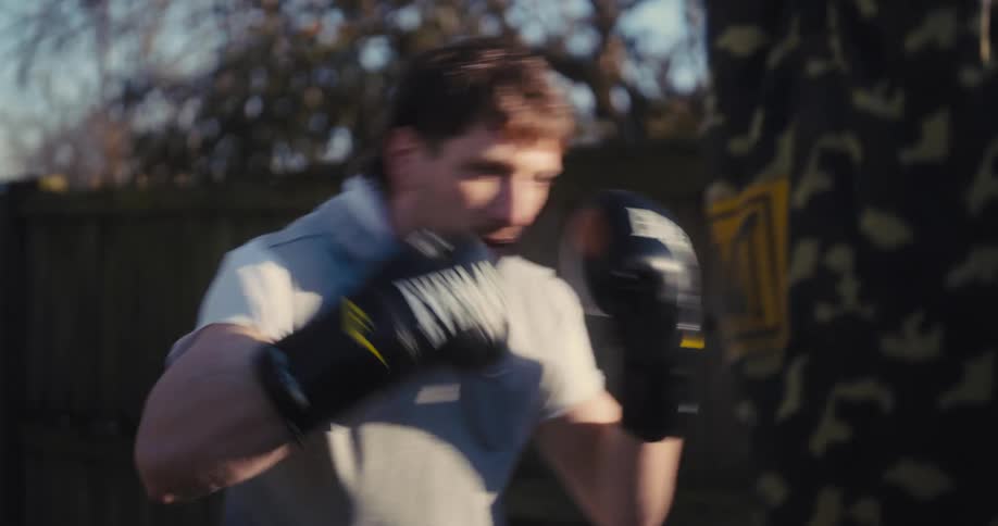 A man is practicing boxing outdoors wearing black everlast gloves and a grey sleeveless hoodiehe is focused on hitting a camouflagepatterned heavy bag while standing in front of a wooden fence