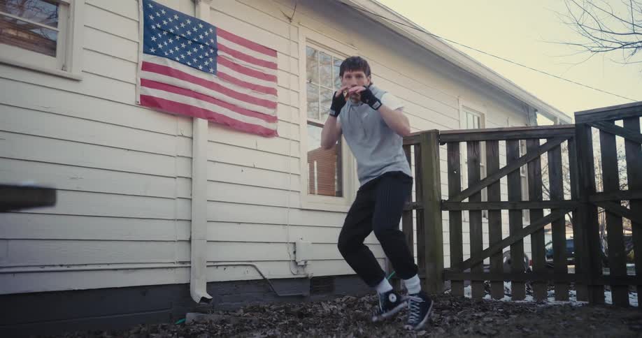 A man wearing a grey sleeveless shirt and black pants practices boxing movements in a backyardan american flag hangs on the white siding of the house behind him as he throws punches toward the camera