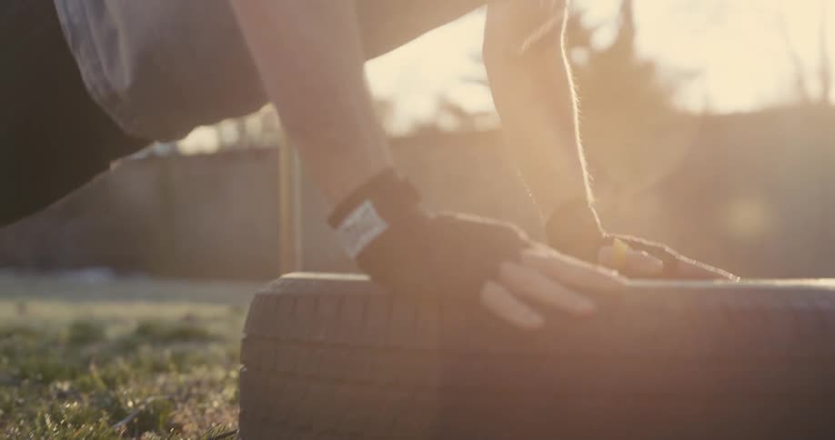 A close view of a man wearing black hand wraps and a grey vest performs push ups on a large tire outdoors as the bright evening sun creates a strong lens flare in the background