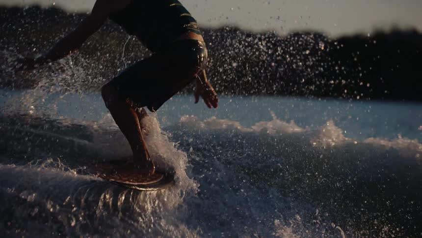 A close view of a man wearing dark shorts and a vest as he balances on a board while surfing through splashing white water on a lake at sunset