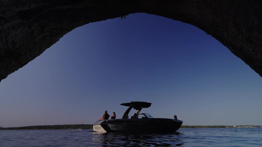 A view from inside a dark cave shows a group of friends on a motorboat floating on a calm lake as one person dives into the blue water under a clear sky
