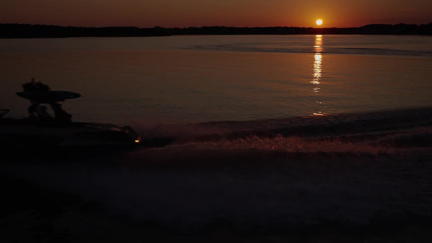 A dark silhouette of a motorboat carrying a group of friends speeds across a calm lake at sunset as the orange sun reflects on the water surface and dark land stretches across the horizon