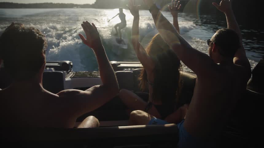 The video captures a group of friends celebrating on a boat while watching a person wakeboard through the sunlit wake the scene is shot from a low angle behind the passengers highlighting the energetic atmosphere as they raise their arms against the glo