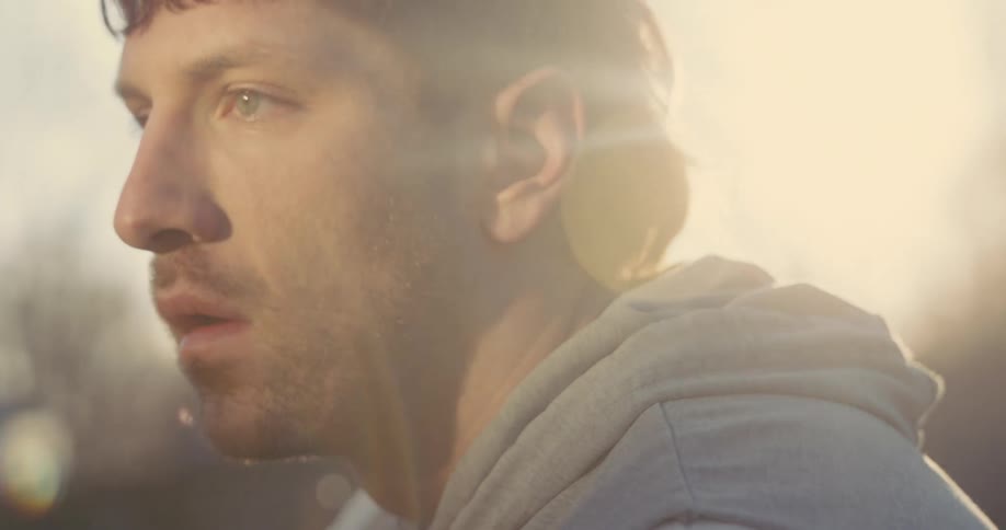 A close view of a man with short dark hair and a light beard looking toward the side with a focused and tired expression while standing outdoors in the warm glow of the evening sun