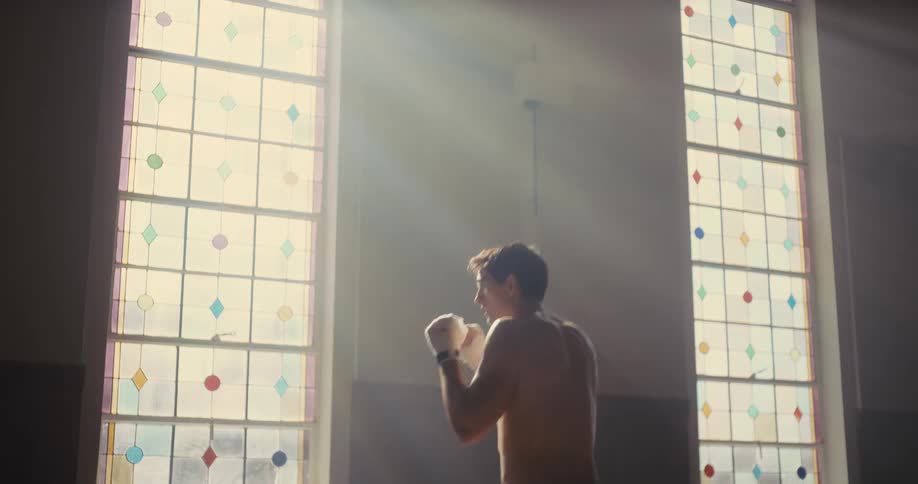 A shirtless man with hand wraps practices shadowboxing in a room illuminated by sunlightthe light streams through two tall stainedglass windows as he throws punches in a steady rhythm