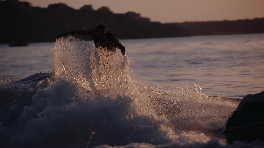 A silhouetted man wake surfs behind a boat during a golden sunset