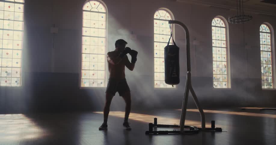 A shirtless man in boxing gloves practices his footwork and punches against a heavy bag in a sunlit cavernous roomatmospheric light streams through large arched windows illuminating the dust and smoke around the athlete as he trains
