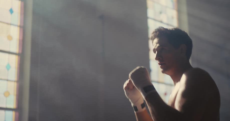 A shirtless man shadowboxes intensely inside a sunlit church sanctuary interior