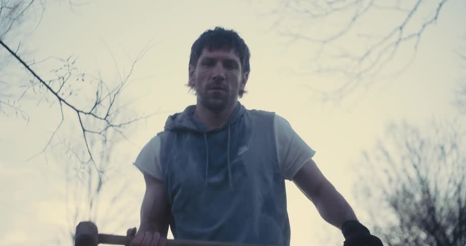 A man wearing a grey hooded vest and a white shirt swings a heavy mallet downward while standing outdoors against a pale sky and bare trees during a workout