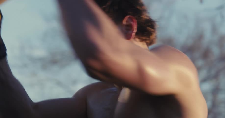 A close view of a shirtless man outdoors as he swings a heavy mallet downward in a repeated motion while training during a clear day