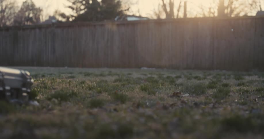 A shirtless man wearing black hand wraps swings a heavy mallet to strike a large tire on the grass in a backyard during a bright day with a clear sky