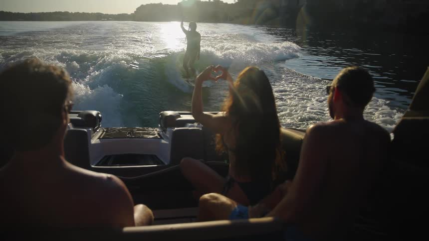 A group of friends watches from a boat as a person wakeboards behind them silhouetted against the bright sunone woman in the boat makes a heart shape with her hands toward the wakeboarder while they cruise across the water at sunset