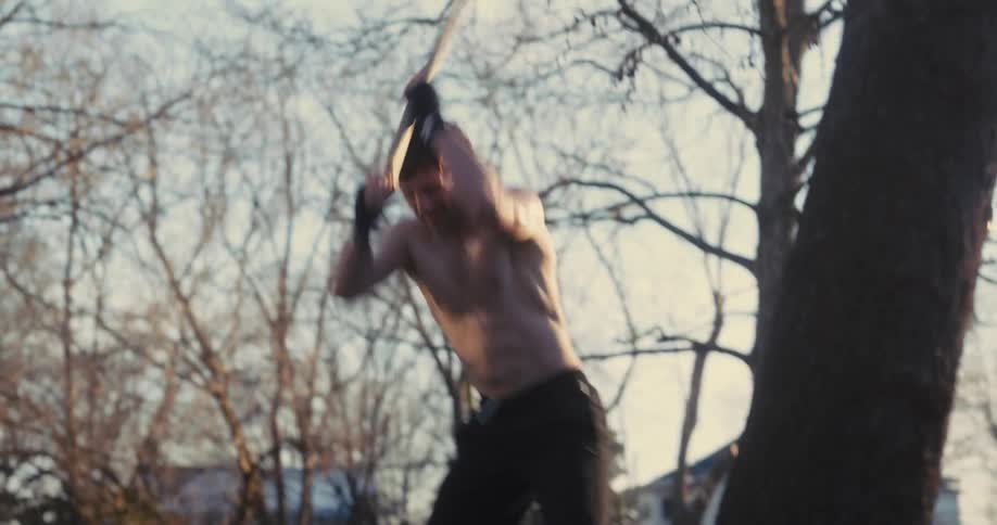 A shirtless man wearing black hand wraps swings a heavy mallet to strike a large tire on the grass in a backyard during a bright day with a clear sky