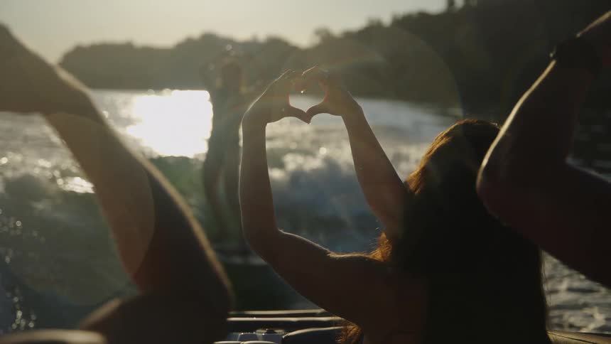 A woman on a boat makes a heart gesture with her hands toward a wakeboarder riding the waves behind them the scene is captured at sunset creating a warm golden glow across the water and silhouettes
