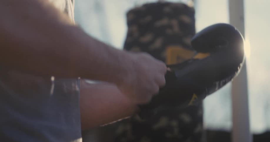 A man prepares for a boxing workout outdoors by carefully putting on and securing his black everlast gloveshe stands in the sunlight near a hanging camouflage punching bag readying himself for training