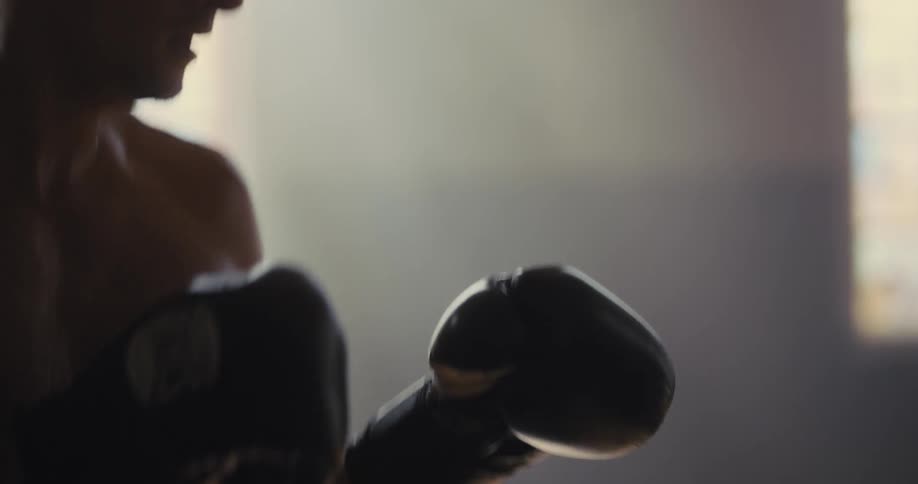 A close view of a shirtless man wearing black boxing gloves as he throws rapid punches toward the camera in a dimly lit indoor space with soft light