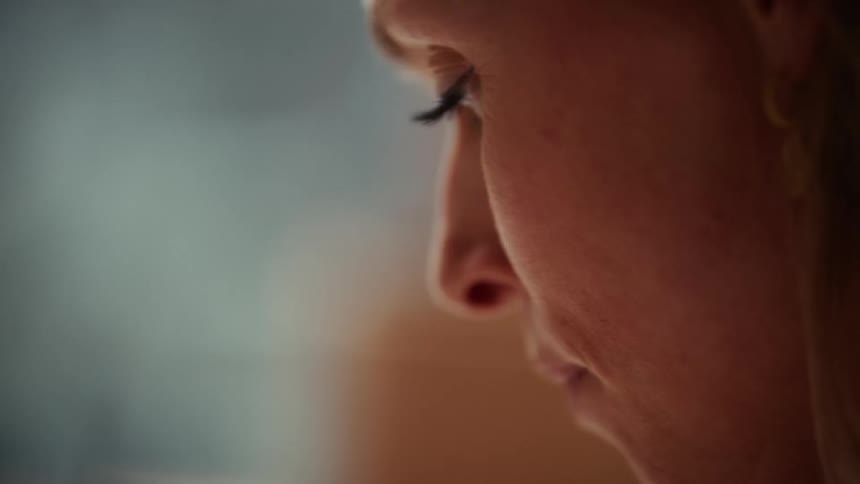 A close view in profile of a woman with light hair looking downward with a neutral expression as soft light illuminates her face in a dimly lit setting