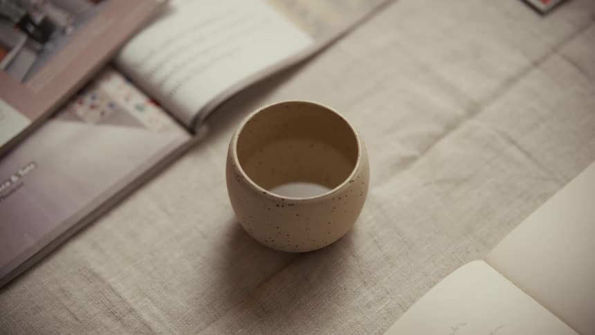 A small speckled ceramic cup sits empty in the center of a light linen tablecloth surrounded by the soft edges of open magazines and books in a bright indoor setting