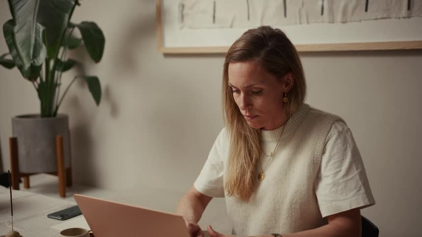 The woman is wearing a dark blue business suit and sitting in a crowded noisy openplan officeshe is actively participating in an energetic video call laughing and gesturing toward the camera