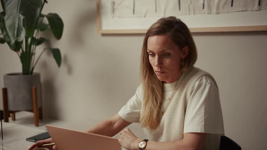 A woman sits at a desk in a welllit room focused on her laptop while holding a pencil she is dressed in a white tshirt and a creamcolored knit vest accessorized with gold jewelry and a watch