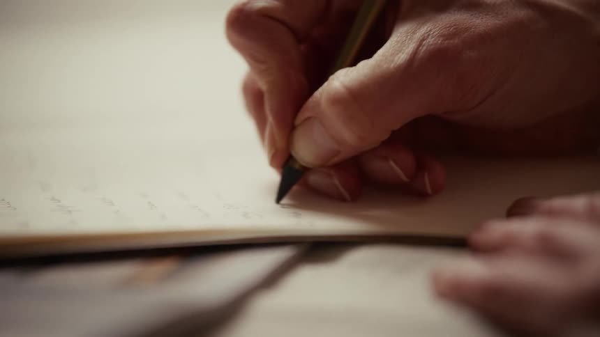 A close view of a hand holding a dark pencil and writing on a piece of cream paper during the day