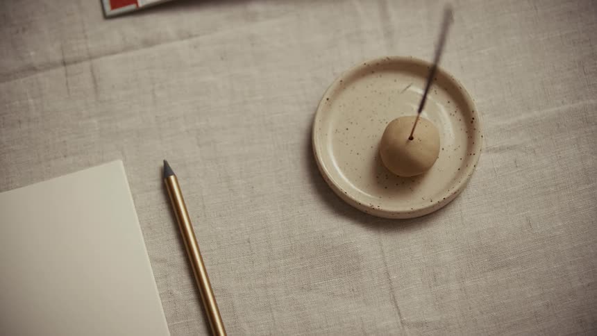 Incense holder with lit stick on ceramic dish beside gold pencil and blank paper