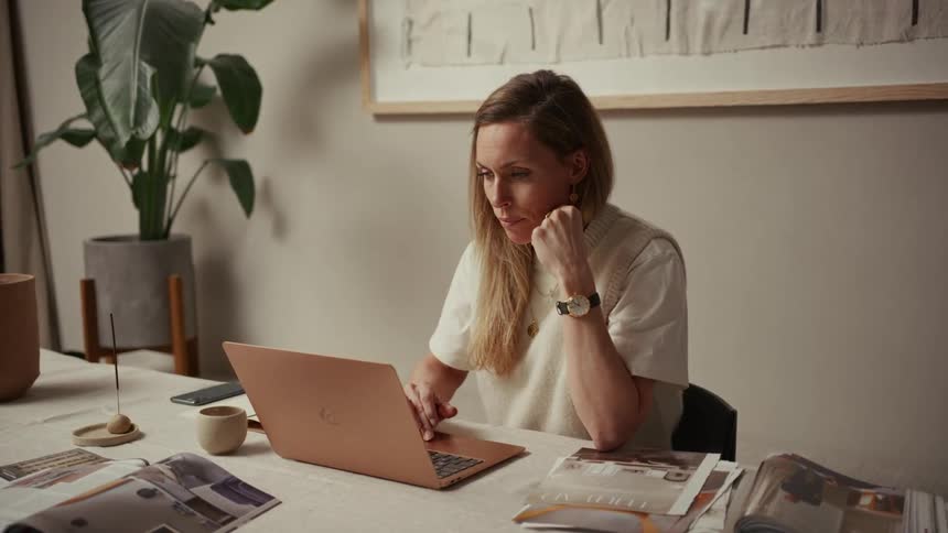 Person works on rose gold laptop at table with magazines and decorative items