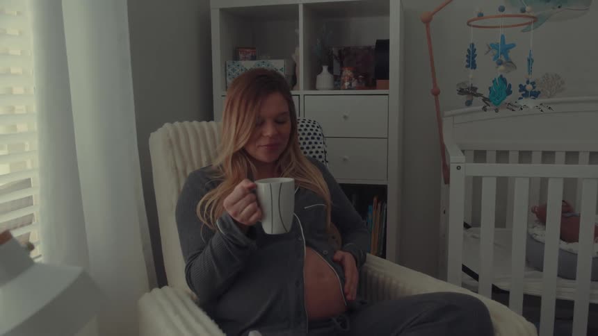 Pregnant woman sitting in an armchair holding a mug next to a crib in a room