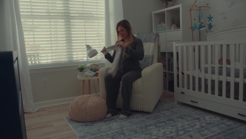 The video shows a woman sitting in a nursery carefully folding a small piece of baby clothing while looking down at it with a smileshe is seated in a lightcolored rocking chair next to a white crib surrounded by typical nursery decor like a patterne
