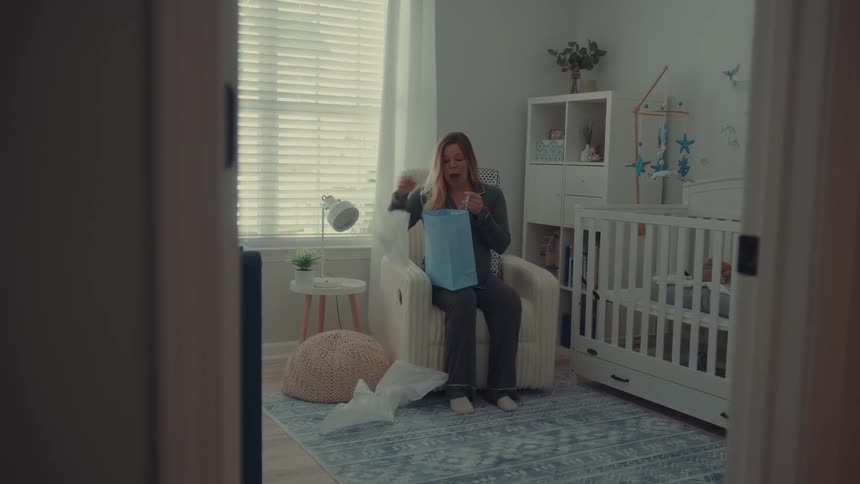 Pregnant woman sitting in a nursery opening a gift next to a crib
