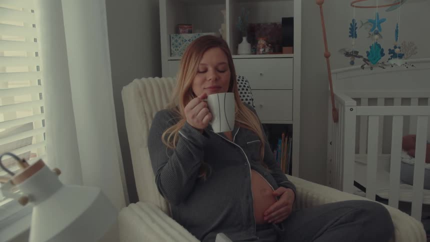 A young pregnant woman sitting beside a baby crib near the window drinking coffee from a mug while rubbing her baby bump