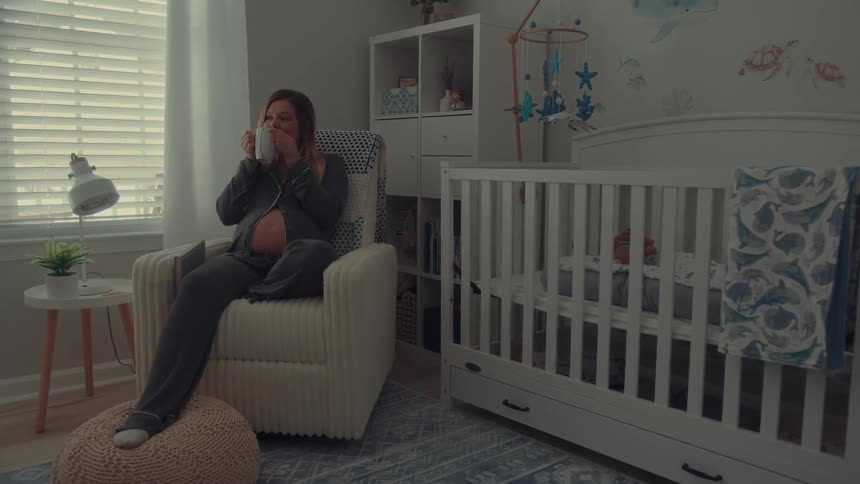 A pregnant woman sits in a rocking chair in a nursery quietly sipping from a mug while looking toward a windowthe room is softly lit and features a white crib oceanthemed wall decals and a storage shelf filled with baby items