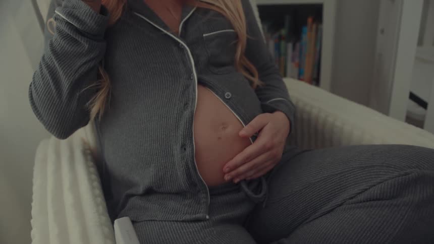 Pregnant woman seated in armchair holds mug and touches belly
