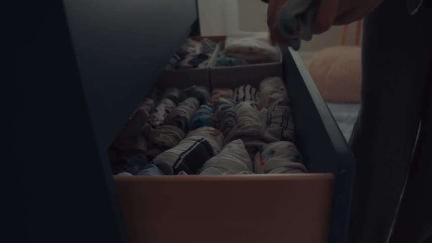 A pregnant woman wearing grey pajamas stands in a nursery as she places clothes into a blue dresser drawer filled with organized baby clothes