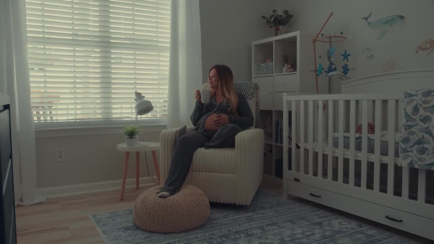 Pregnant woman sitting in an armchair drinking a hot beverage in a room with a crib and a lit window