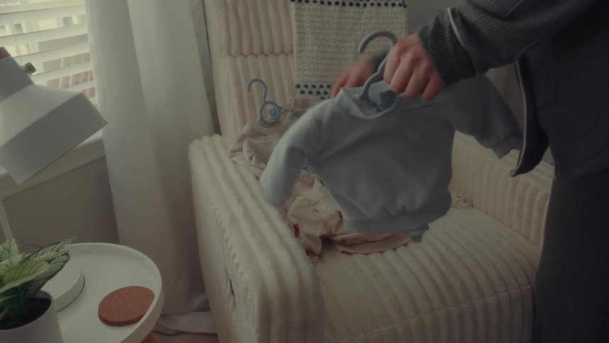 A person picks up small baby sweaters on a hanger from a pile of baby clothes sitting on a cream colored rocking chair in a nursery during the day