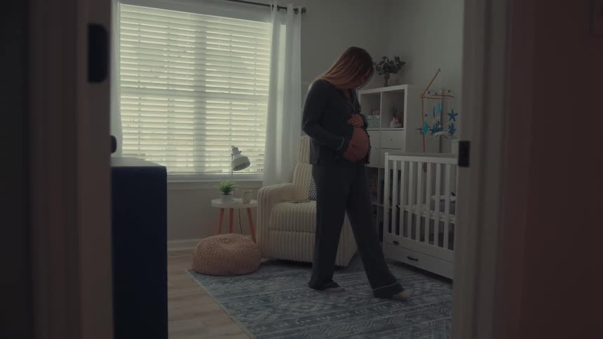 A pregnant woman wearing grey pajamas stands in a decorated nursery and gently rubs her stomach while looking down near a white crib and a cream colored rocking chair