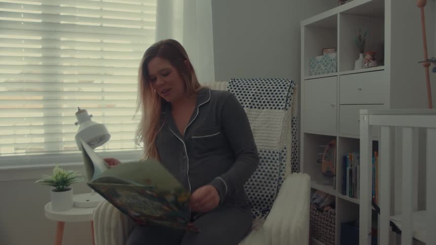 A pregnant woman wearing pajamas sitting in an armchair and smiling as she reads a baby storybook