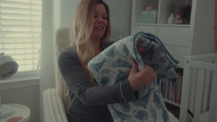 A smiling pregnant woman sits in a nursery chair looking down at a blue baby blanket patterned with whalesshe carefully folds the blanket while sitting next to a white crib in a brightly lit room
