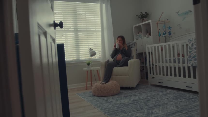 A pregnant woman sits in a white rocking chair within a nursery holding a mug and resting her feet on a poufthe room features a white crib a blue patterned rug and whalethemed wall art near a window with closed blinds
