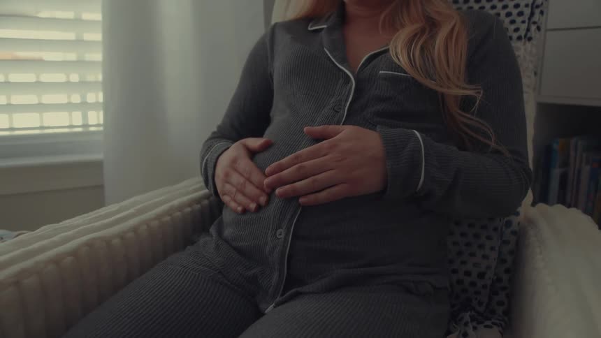 A pregnant woman wearing gray pajamas sits in a rocking chair and gently rubs her stomach with both hands in a dimly lit room with a window and bookshelf in the background